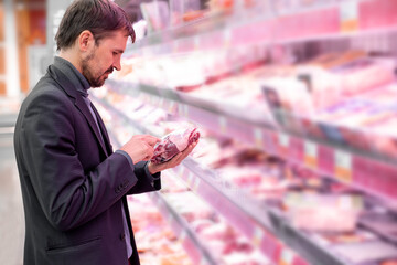 A bearded man reads information about the product, stands near the refrigerator in the supermarket in the meat department