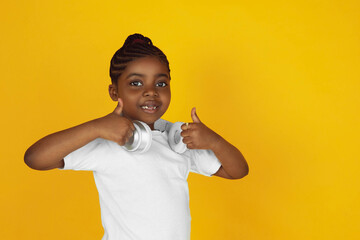 Thumb up, putting on headphones. Little african-american girl's portrait on yellow studio background. Cheerful kid. Concept of human emotions, facial expression, sales, ad. Copyspace. Looks cute.