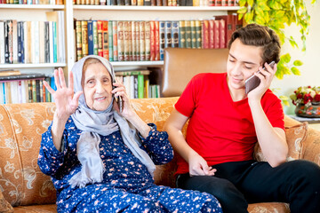 Wall Mural - Happy arabic muslim grand mother and her son sitting together on sofa and using mobiles