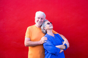 couple of seniors having fun together and smiling - mature man looking at the camera and hugging his wife - red background - portrait sand close up