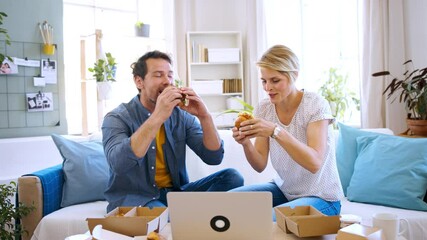 Wall Mural - Happy couple sitting on sofa indoors at home, eating hamburgers.