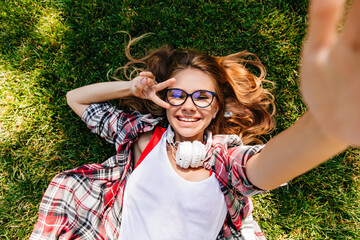 Wall Mural - Pleasant caucasian lady making selfie while lying on lawn. Laughing good-looking girl expressing happiness in summer weekend.