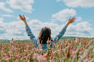 Back view of young woman in pink flower field with arms up towards the blue sky