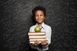 © millaf - Smart black child student smiling and holding books and green apple against blackboard background with science and maths formulas