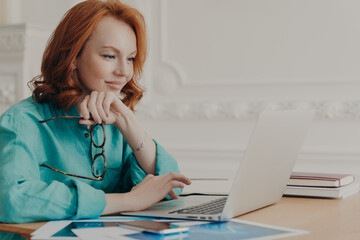 Wall Mural - Busy redhead woman entrepreneur works on modern laptop, holds spectacles, poses in coworking space, reads email, received good business news, does remote job from home, enjoys informal chat.