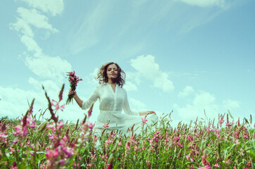 Front view portrait of young woman in motion in a long white dress walking with a wild flower bouquet in a blossoming flower field holding dress with hand