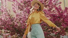 Elegant Fashionable Woman Wearing Straw Hat, Yellow Sunglasses, Silk Blouse, Light Blue Trousers, With White Round Wicker Shoulder Rotang Bag Walking In Street, Near Blooming Trees