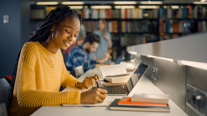 university library: gifted black girl uses laptop, writes notes for the paper, essay, study for clas