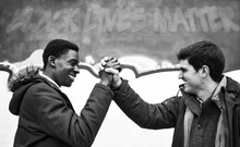 Black And White Image Of The Joined Hands Of Two Young Men Of Different Ethnicity Smiling At Each Other In Front Of A Blurred Wall With The Slogan "black Lives Matter". Stop Racism Campaign