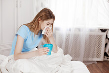A Sick Girl Is Sitting Under A Blanket On A Bed In A Bedroom, Holding A Paper Handkerchief, Blowing Her Nose, Experiencing Relief Of Nasal Congestion And Drinking Hot Medicine.
