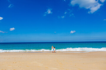 Wall Mural - Young beautiful mother with her charming daughter in white clothes walk on a sandy beach near the water