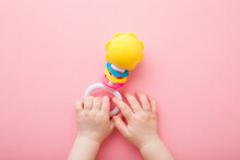Baby Girl Hands Playing With Colorful Rattle On Light Pink Floor Background. Pastel Color. Closeup. Toys Of Development For Infant. Point Of View Shot. Top Down View.