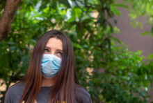 Attractive Brunette Wearing A Light Blue Surgical Mask Looking Into The Distance With A Worried Unsure Look On Her Face. Sunlight Hitting Her On A Sunny Day Outside In The Park