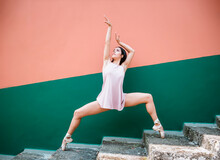 Full Body Young Brunette Ballerina In Pointe Shoes Standing On Tip Of Toes On Weathered Steps And Raising Arms While Dancing Against Striped Wall On Street
