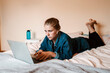© JAKE JAKAB/ADDICTIVE STOCK - Thoughtful focused female in true wireless earbuds and casual clothes lying down on bed using laptop against beige wall in light apartment