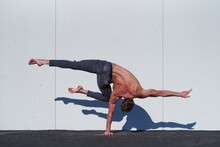 Back View Of Faceless Muscular Shirtless Acrobat Standing On One Hand While Stretching One Leg Perpendicular To Floor And Other Bending At Knee During Training Against White Wall