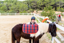 Little Jockey In Protective Helmet Adjusting Stirrup On Saddle Before Riding Skewbald Pony In Equestrian School