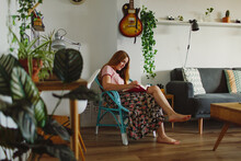 Side View Of Barefoot Female Sitting On Chair While Reading A Book In Cozy Living Room At Home