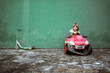 © Rafa Cortes/ADDICTIVE STOCK - Cute little girl in casual clothes looking at camera and driving red electric vehicle with broken bumper against shabby green wall near kick scooter