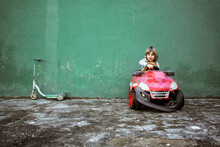 Cute Little Girl In Casual Clothes Looking At Camera And Driving Red Electric Vehicle With Broken Bumper Against Shabby Green Wall Near Kick Scooter