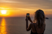 Woman Hands Holding Mobile Phone At Sunset. Young Curly Hair Woman Taking Photos With Her Cell Phone In A Beautiful Amazing Sunset Over Sea. Taking A Picture On A Smartphone During A Vacation
