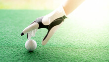 Female Hand Wearing White Professional Gloves With The Left Hand Putting Golfball On The Rubber Tee Grass Course.