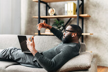 Serious African-American Guy In Glasses With Airpods Earphones Lays On The Sofa In Stylish Living Room And Using Trendy Laptop For Video Call. Remote Work Concept