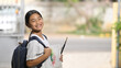 © Prathankarnpap - A schoolgirl is holding painting equipment and carrying a school bag while standing and waiting for a school bus.