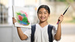 © Prathankarnpap - A schoolgirl is holding painting equipment and carrying a school bag while standing and waiting for a school bus.