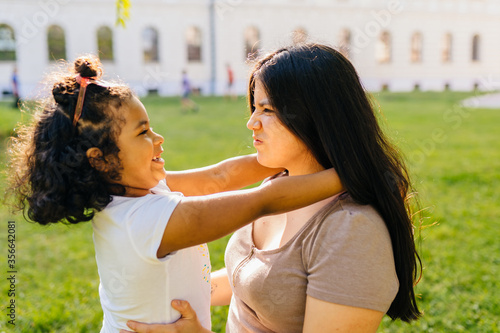 Fotomural Cutie curly mexican dark skinned preschooler girl playing, hugging her latin mother outdoor in european city park