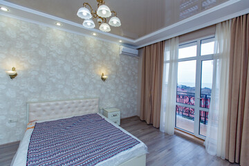 Room in bright colors with a new renovation. A large white bed and a white chest of drawers opposite the bed.Floor-length window with beige curtains