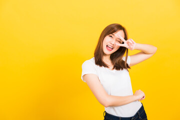 Asian happy portrait beautiful cute young woman teen smile standing wear t-shirt showing finger making v-sign symbol near eye  looking to camera isolated, studio shot yellow background with copy space