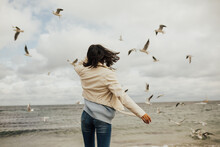 Beautiful Girl Standing At The Sea With Seagulls Around. Happy Young Woman Looking At Flying Seagulls At The Sea Beach. Girl Traveler. Concept Of Freedom, Travel. Lifestyle Moment. Copy Space.