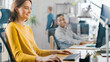 © Gorodenkoff - Beautiful and Creative Young Woman Sitting at Her Desk Using Laptop Computer. She Chats, Smiles and Jokes with Her Friendly Colleague. Office where Diverse Team of Young Professionals Work.