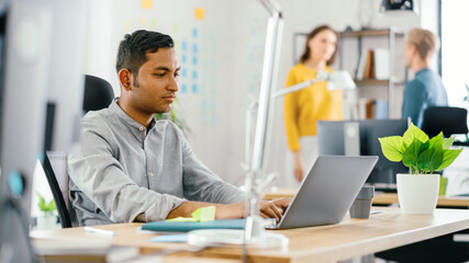 Smart and Handsome Indian Office Worker Sitting at His Desk works on a Laptop. In the Background Modern Office with Diverse Team of Young Professionals Working.