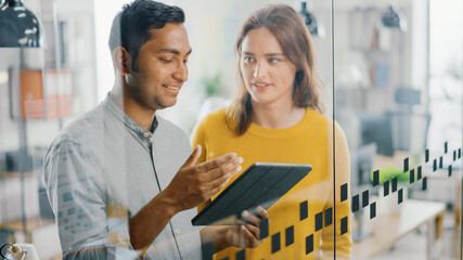 Portrait of Handsome Indian Man Showing Touch Screen Digital Tablet Computer To His Beautiful Female Team Member, They Discuss Project Details. Successful Young People Working in Diverse Office.