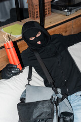 Robber in balaclava holding dynamite near bag with laptop on couch