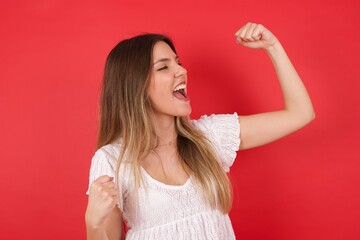 Attractive young caucasian woman celebrating a victory punching the air with her fists and a beaming toothy smile over a white studio background with copy space.