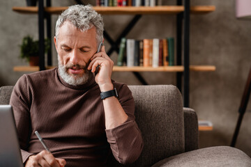 Senior man in casual talking on mobile phone and writting notes while working at home using laptop