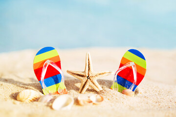 Travel background with bright flip-flops and starfish on the sand against the background of the blue sea, closeup, soft focus. Beach summer vacation. 