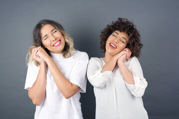 Sticker - Dreamy young female with pleasant expression, closes eyes, keeps hands crossed near face, thinks about something pleasant, poses against gray background.