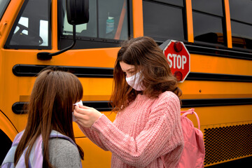 Children with face masks by school bus. Two girls help each other with facemasks while standing by a school bus. Education, health, safety, back to school, coronavirus, and school concepts.