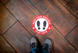 © F Armstrong Photo - Floor sign on wood floor at business stating Social Distancing Stay Safe with shoes as graphic . A person’s legs and shoes can be seen nearby