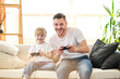 © Pavel Shcherbakov - Father and daughter. Handsome young man and his child little cute girl are playing game console, looking at camera and smiling while sitting on couch at home. Father's day.