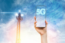 A Woman's Hand Holds A Smartphone Against A Blue Sky With A 5G Telecommunications Tower. Concept Of Modern Technology And Telecommunications