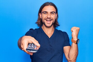 Young handsome man holding television remote control screaming proud, celebrating victory and success very excited with raised arms