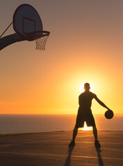 Basketball player dribbling a ball alone at sunset on outdoor court with a beach background. Silhouette. 