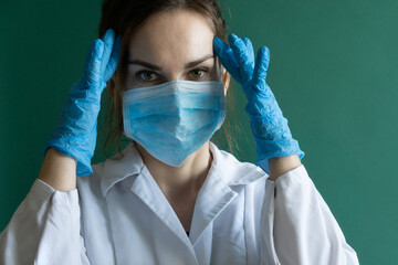 A woman in a white overall with a mask on her face and surgical gloves on hands. Portrait of a young European doctor staring off into space.