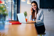 Skilled female copywriter talking on mobile discussing details of task updating application on laptop