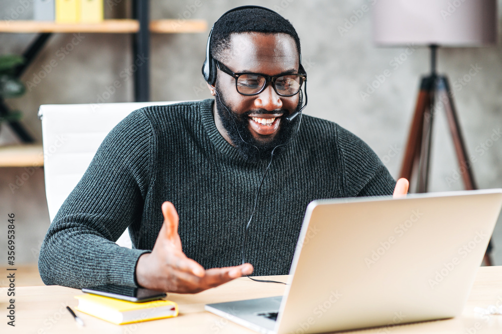 Smiling African-American guy uses a handsfree headset to talk online at his workplace, black confident man in glasses sits at the office desk and looks at laptop screen - obrazy, fototapety, plakaty 